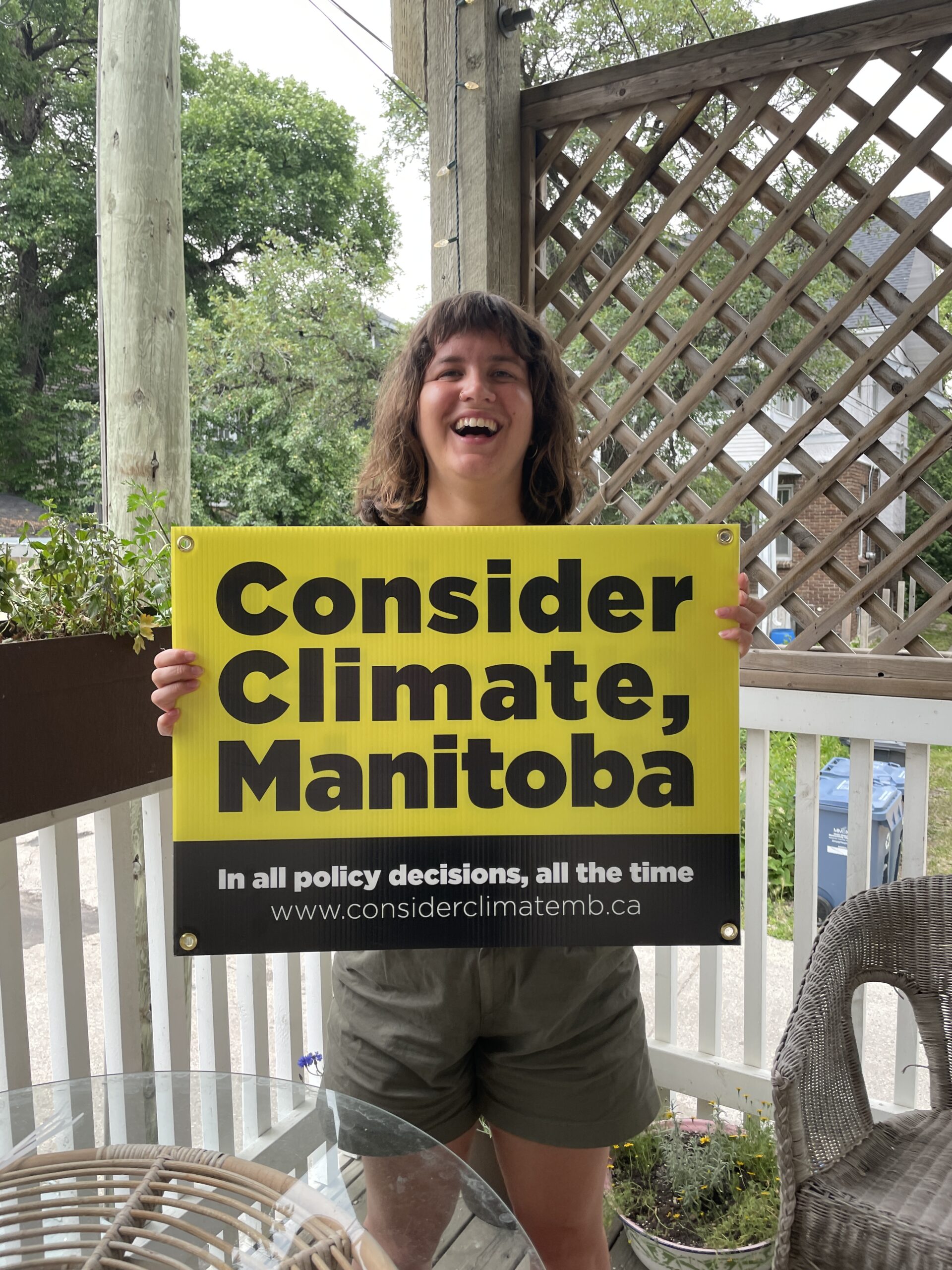 Nat, a young female adult is standing on her porch smiling while holding up a Consider Climate, Manitoba sign