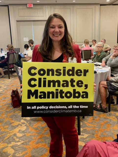 Sherri Rollins, City of Winnipeg counsellor is standing in a room full of people smiling at the camera and holding up a Consider Climate, Manitoba sign. 