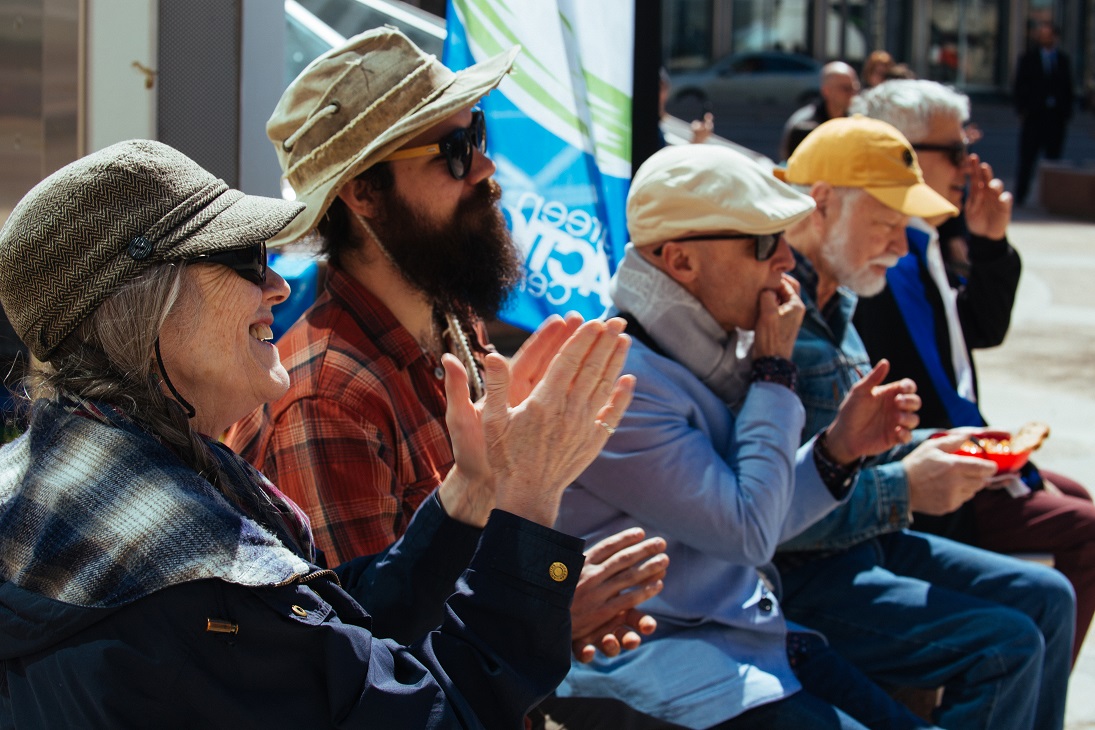 There are 5 people sitting on a bench at an event clapping. Four of them appear to be seniors, and one of them is a younger man with a hat and long beard. The man in the middle is whistling. 