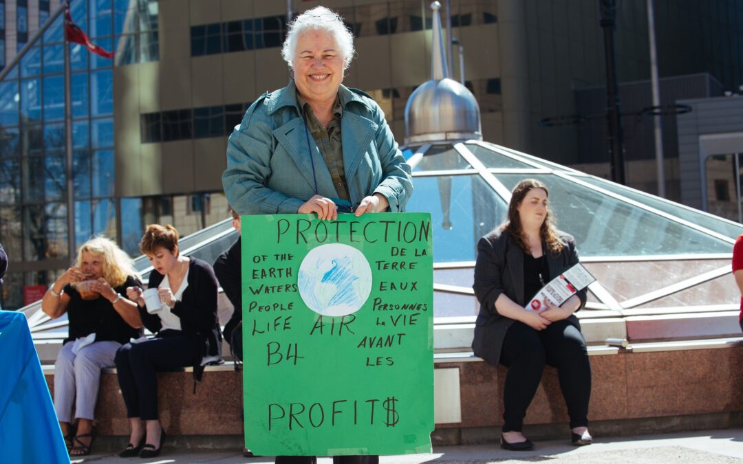 A senior is smiling at the camera at an outdoor event. They are holding up a green poster that says, "Protection of the earth, waters, people, life, air B4 PROFIT$". There's an image of earth, and beside that the sign also says, "Protection de la terre eaux personnes la vie avant les profit$"